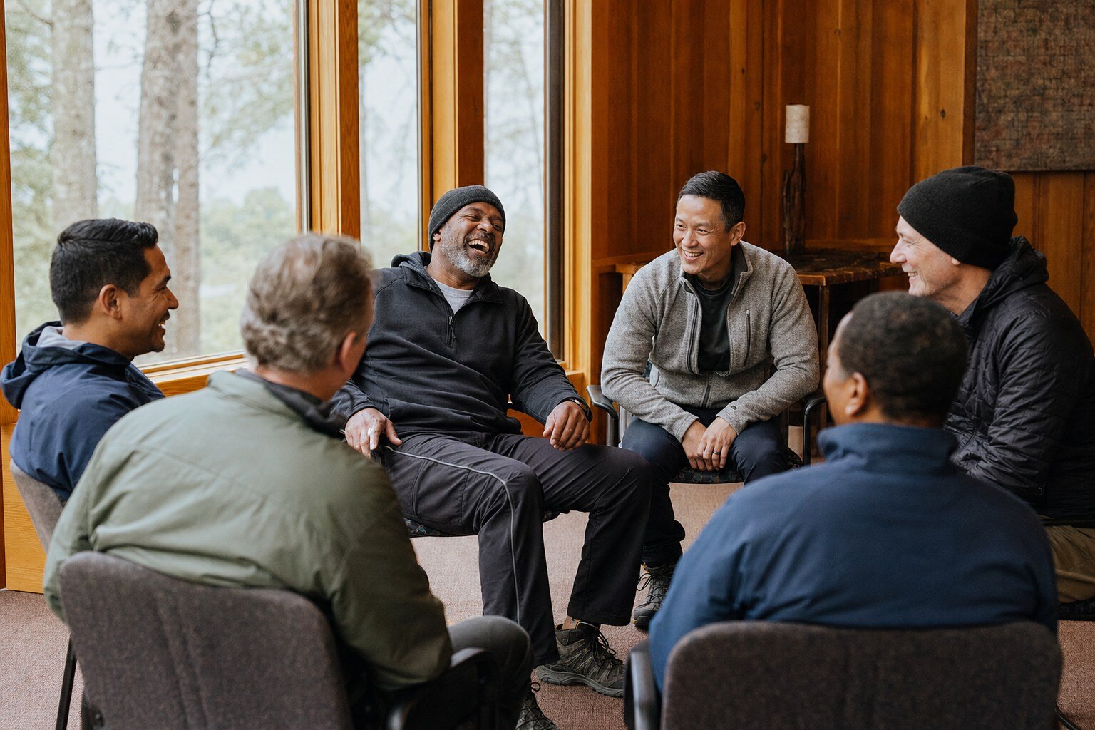 Six longtime friends laughing together in a lodge by tall windows