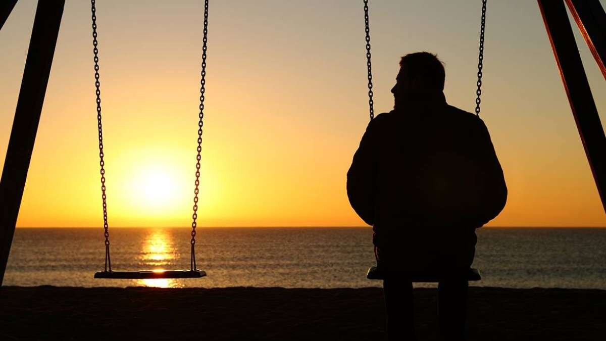 Man sitting alone on a swing at sunset by the ocean, empty swing beside him
